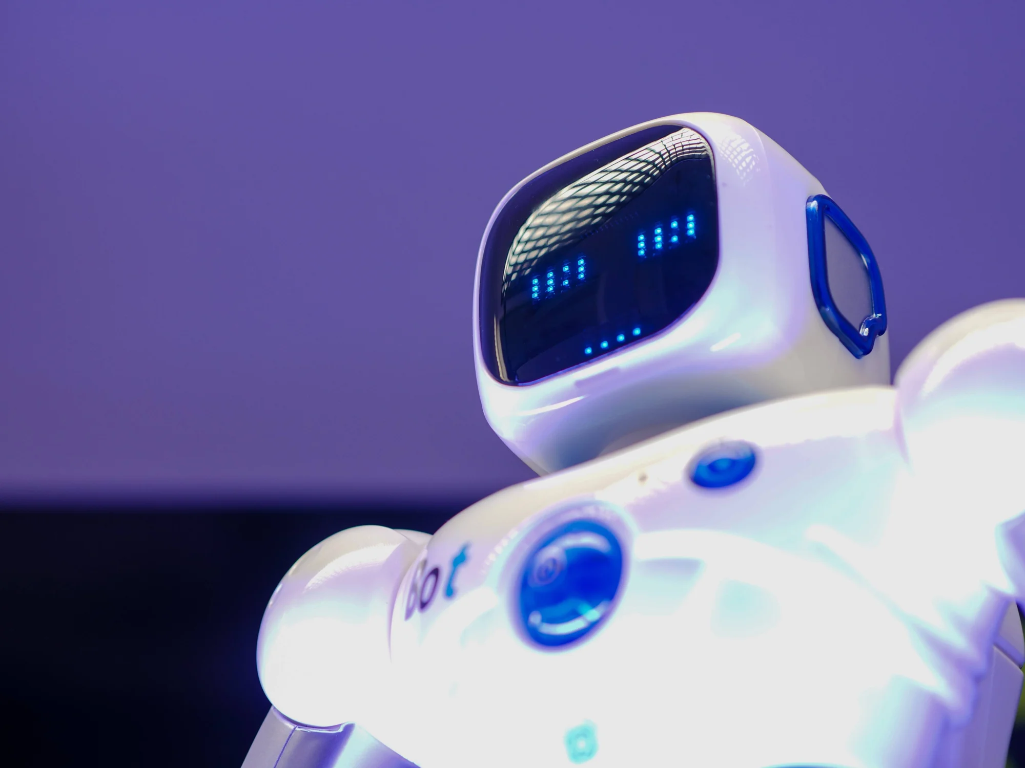 Robot de juguete blanco con pantalla LED azul en la cara y detalles en azul, fotografiado sobre fondo violeta.