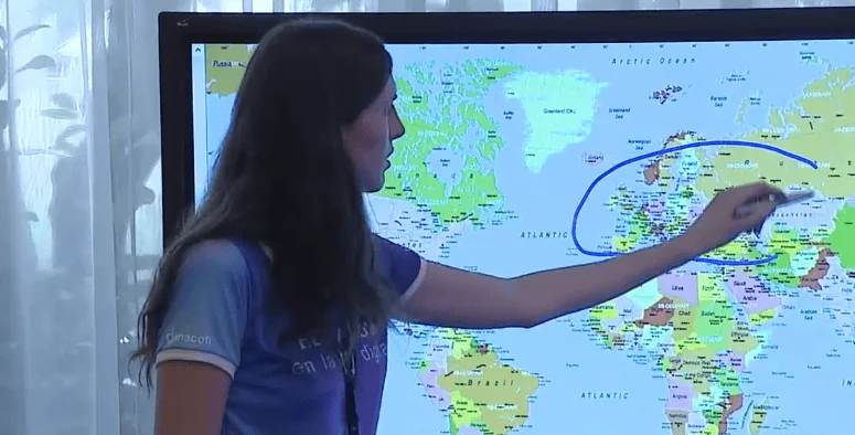 Una mujer señala con el dedo una región de Europa en un mapa mundial proyectado en una pantalla, con una línea azul trazada sobre el océano Atlántico.