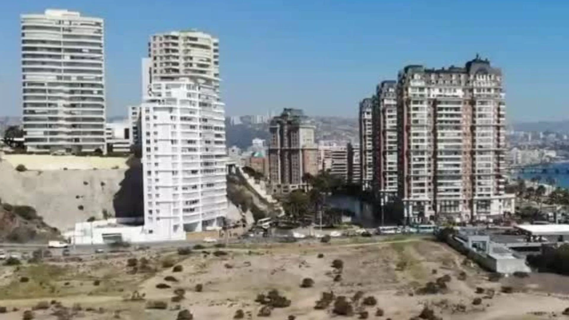 Vista aérea de edificios de gran altura junto a terreno árido en una ciudad costera, con el mar al fondo.