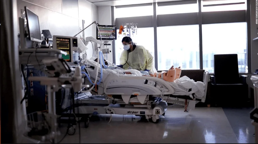 Personal médico con equipo de protección atiende a un paciente en cama hospitalaria rodeada de monitores y equipos médicos.