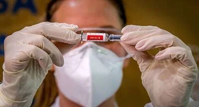 Trabajadora de salud con mascarilla sostiene un vial etiquetado COVID-19 entre sus manos enguantadas.