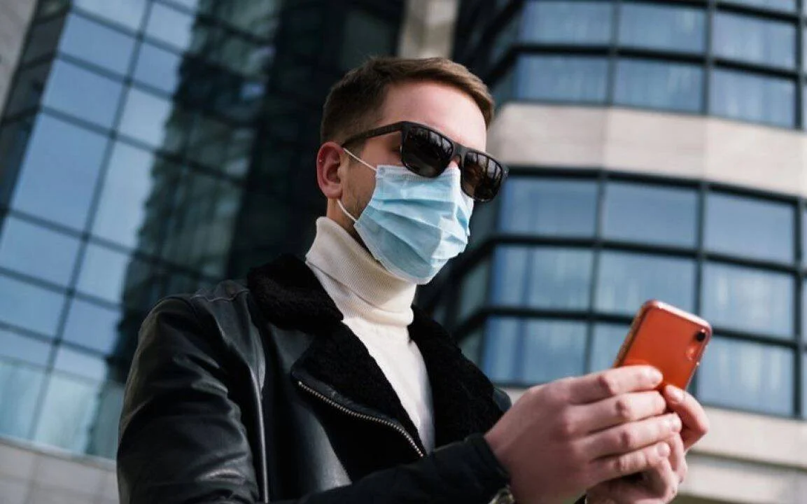 Hombre con mascarilla quirúrgica y gafas de sol revisa su teléfono móvil frente a un edificio de vidrio.