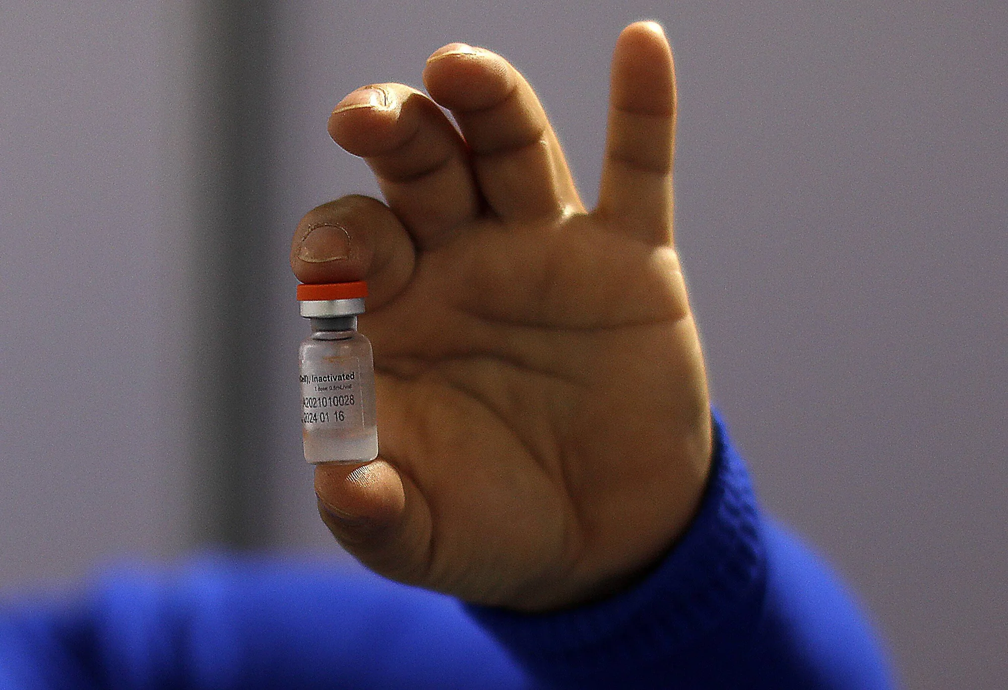 Una mano con guante azul sostiene un pequeño vial de vacuna inactivada con tapa roja.