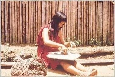 Mujer con vestido rojo sentada en el suelo trabajando con sus manos sobre una pequeña mesa, junto a una pared de cañas.