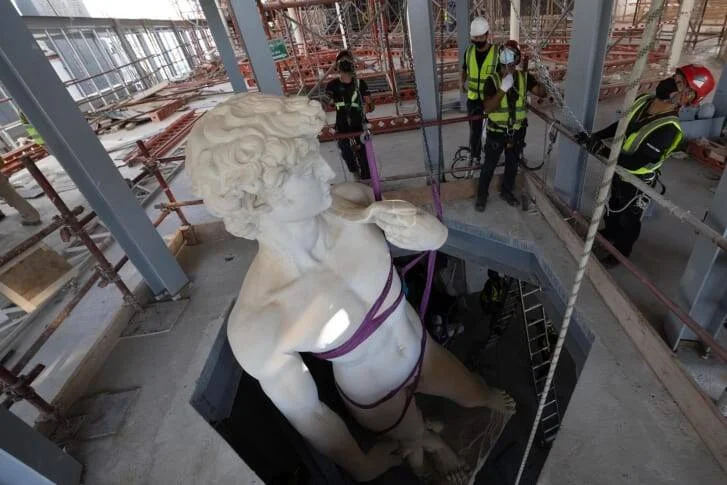 Trabajadores instalan con correas una gran escultura de figura masculina clásica en blanco dentro de un edificio en construcción con andamios.