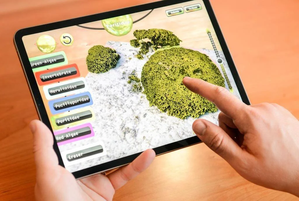 Manos sosteniendo una tablet con una app interactiva para identificar especies de coral y algas en un arrecife simulado en 3D.