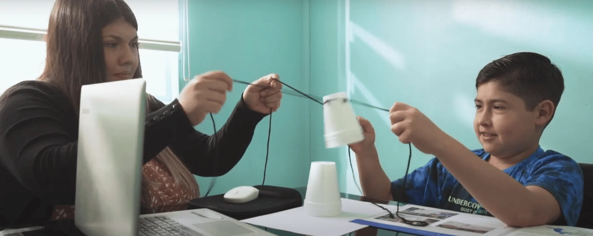 Una maestra y un niño realizan un experimento escolar con vasos de plástico unidos por un hilo, simulando un teléfono casero.