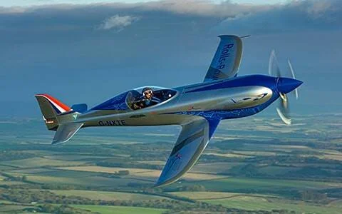 Avión eléctrico experimental azul y plateado de Rolls-Royce volando sobre campos verdes con un piloto en cabina abierta.