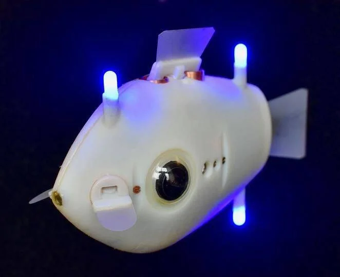 Robot submarino con forma de pez blanco equipado con luces LED azules y cámara frontal, sobre fondo oscuro.