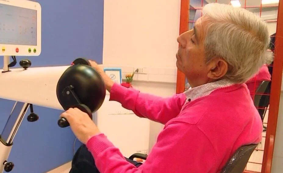 Hombre mayor con suéter rosa opera un equipo de rehabilitación física frente a una pantalla táctil en un centro de salud.