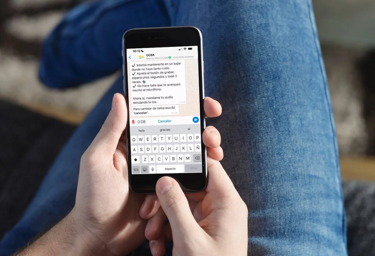 Manos sosteniendo un iPhone con una conversación de WhatsApp del Gobierno de Buenos Aires con instrucciones para grabar un audio de tos.