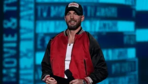 Hombre con chaqueta bomber roja y negra y gorra de NASA sonríe en el escenario de una ceremonia de premios de cine y televisión.