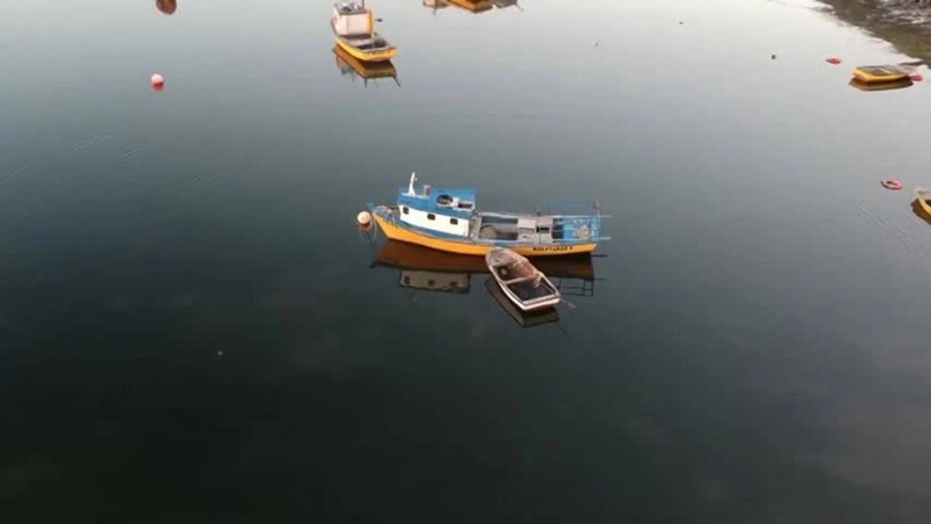 Vista aérea de una embarcación pesquera amarilla y azul anclada en aguas calmas, rodeada de otras pequeñas embarcaciones.