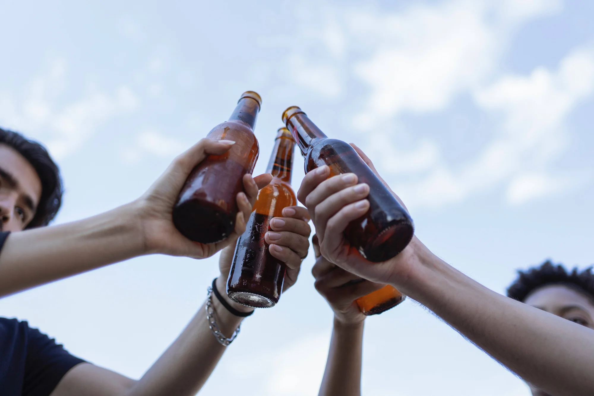 Varias personas chocan botellas de cerveza al aire libre en un brindis, fotografiado desde abajo con el cielo de fondo.
