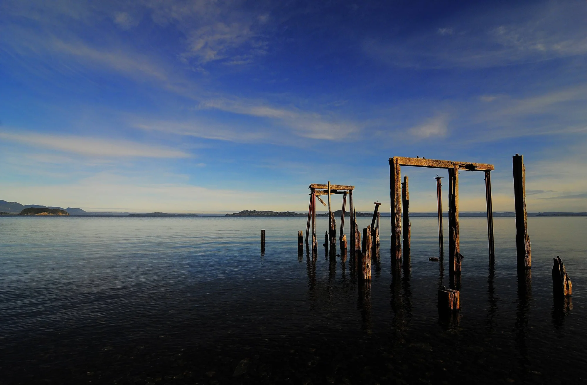 Pilotes y estructura de madera deteriorada de un muelle en ruinas emergen de aguas calmas bajo un cielo azul con nubes.