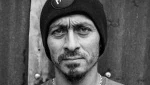 Hombre adulto con gorro negro y collar de cadena mira fijamente a cámara en retrato en blanco y negro.