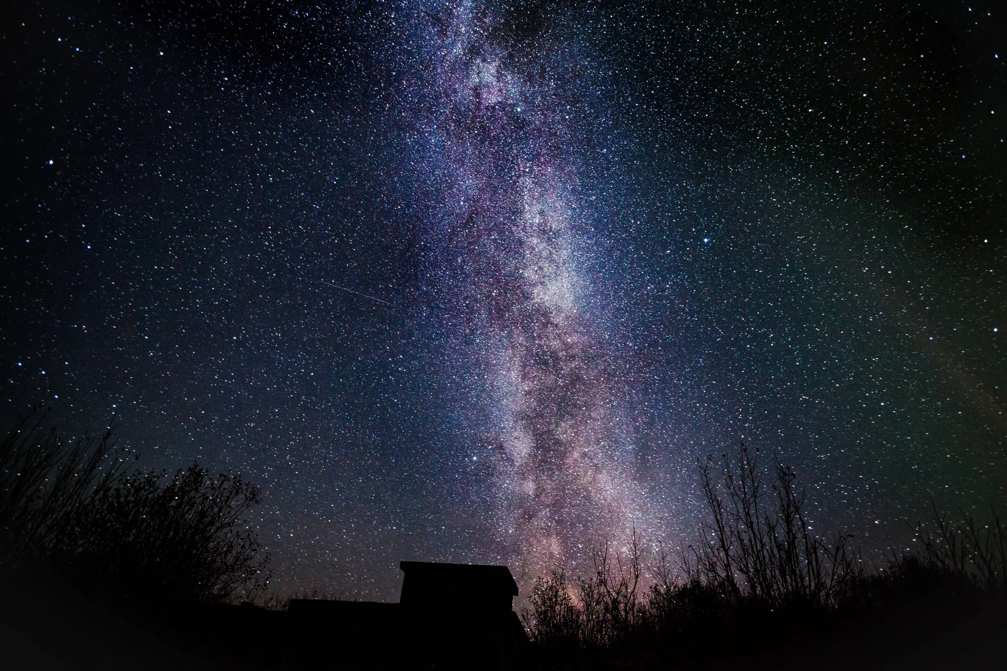 La Vía Láctea se eleva verticalmente en el cielo nocturno estrellado sobre la silueta oscura de una pequeña cabaña y árboles.