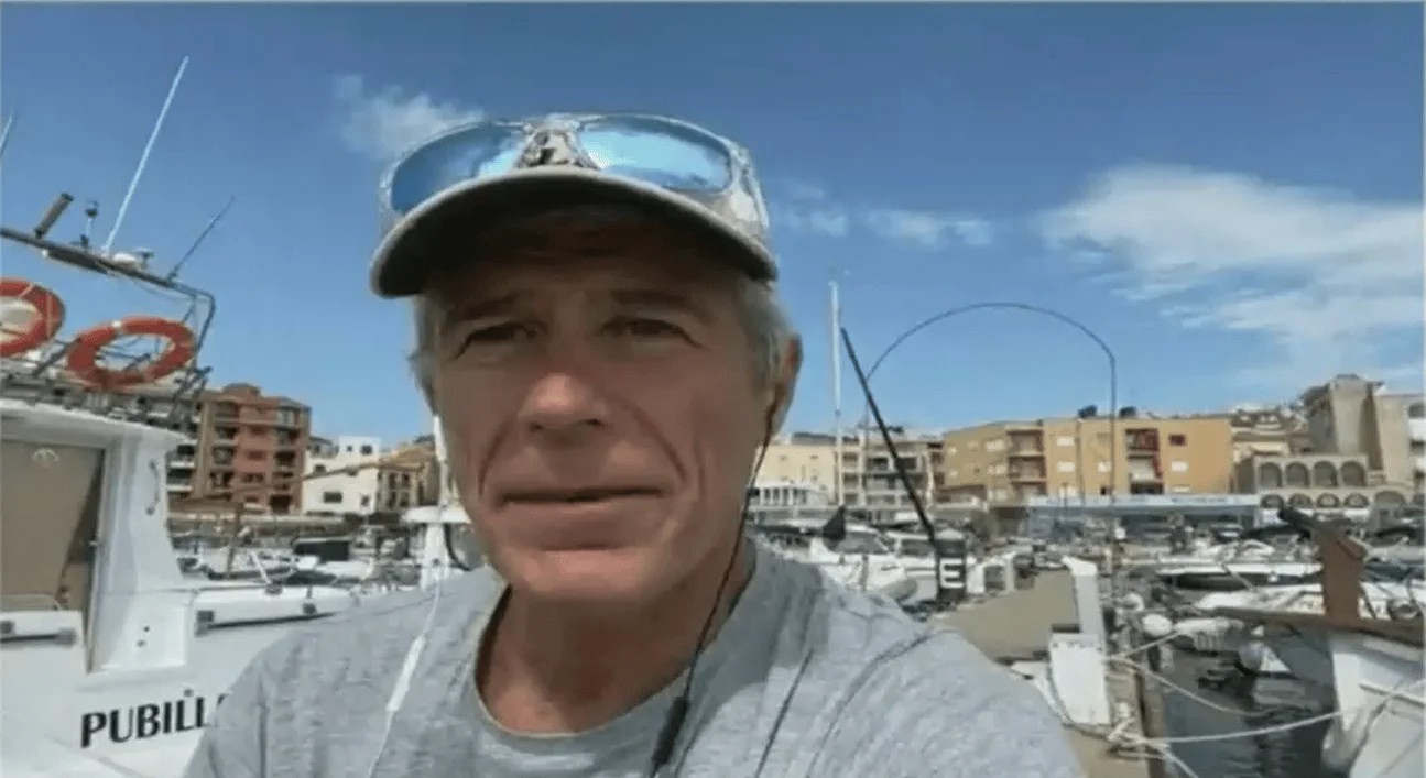 Hombre de mediana edad con gorra y gafas de sol en la cabeza, frente a un puerto deportivo con embarcaciones y edificios al fondo.