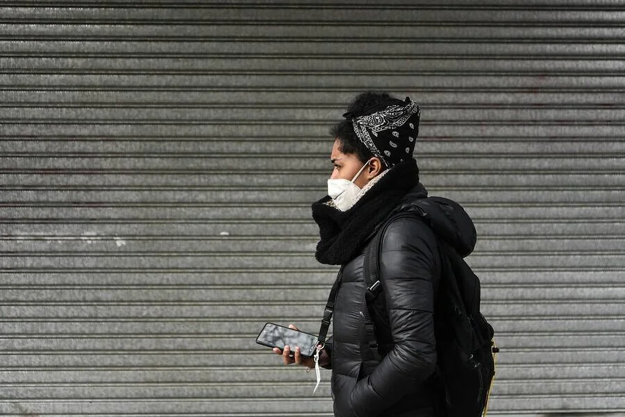 Persona abrigada con mascarilla sanitaria, pañuelo en la cabeza y mochila camina sosteniendo un teléfono frente a una persiana metálica.