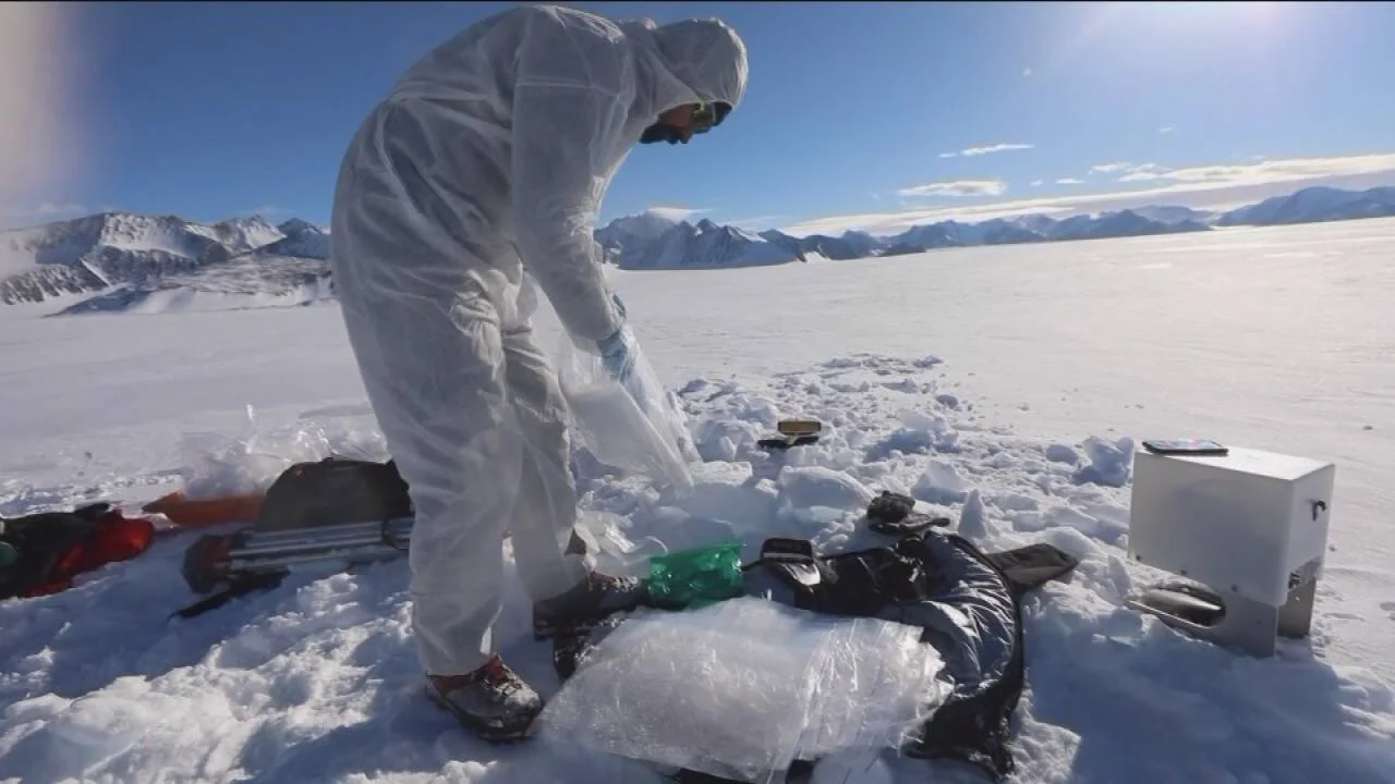 Investigador con traje blanco de protección manipula bloques de hielo en un glaciar nevado rodeado de montañas.