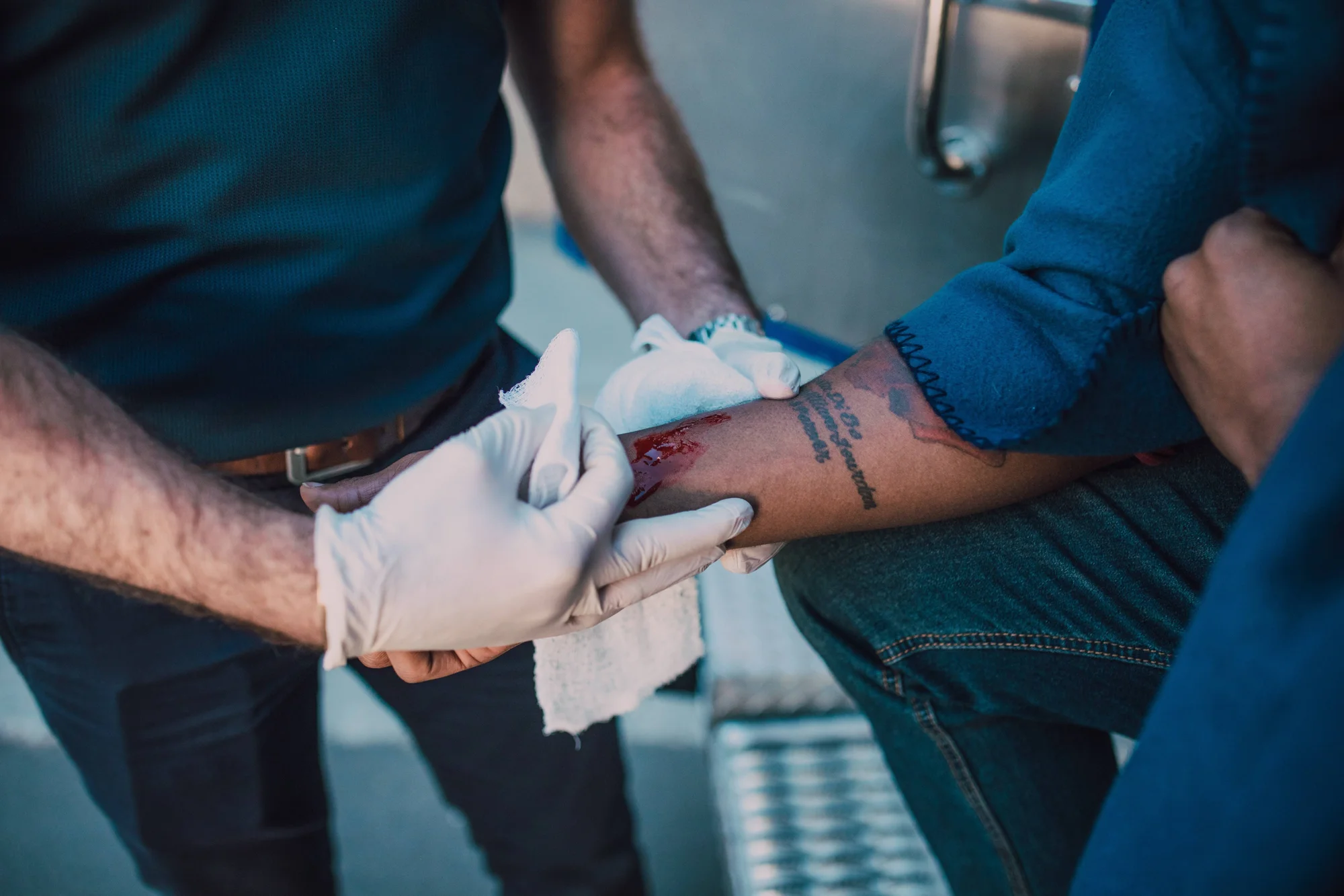 Socorrista con guantes de látex aplica gasa sobre una herida sangrante en el antebrazo tatuado de una persona sentada.