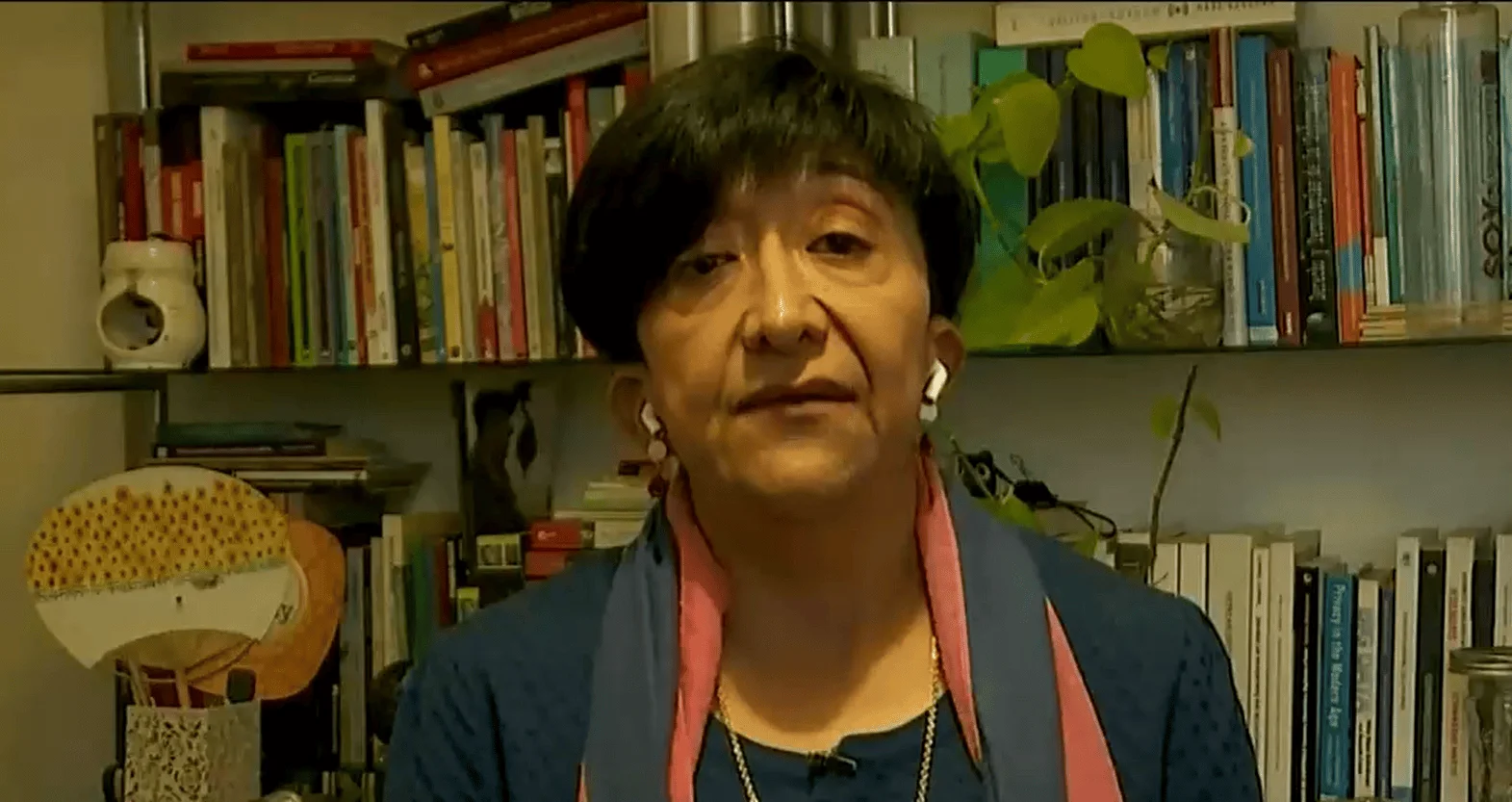 Mujer de mediana edad con auriculares inalámbricos habla frente a una estantería llena de libros y plantas.