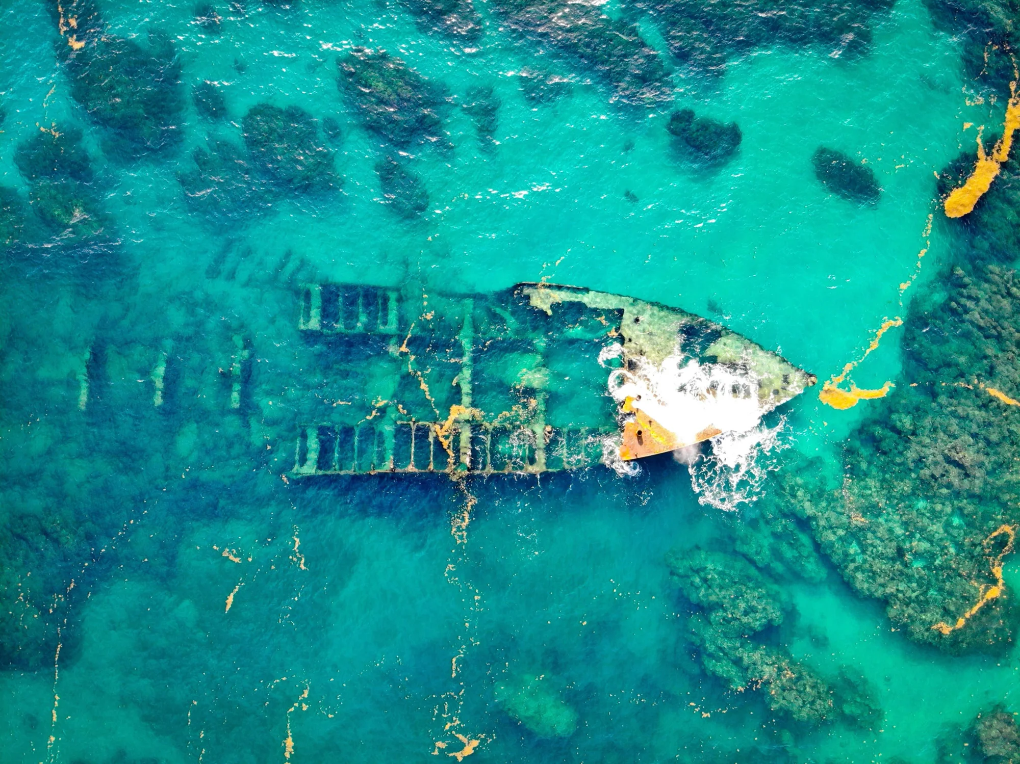 Vista aérea de un barco naufragado semisumergido en aguas turquesas poco profundas con arrecifes de coral visibles.