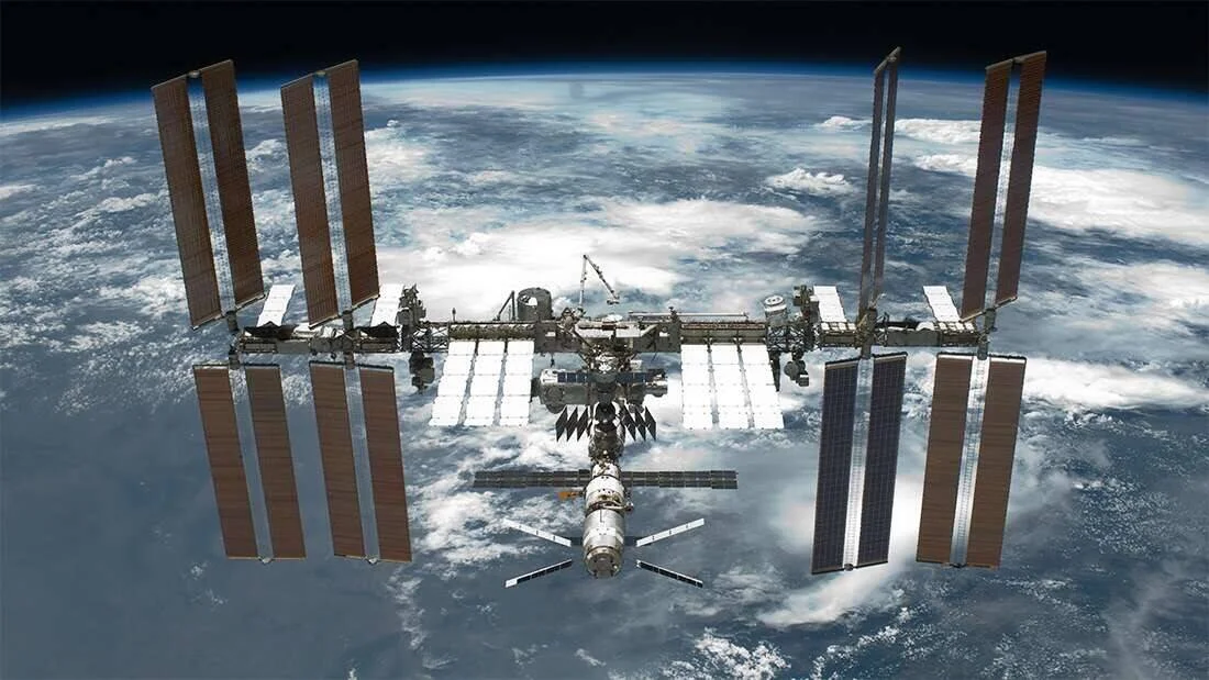 La Estación Espacial Internacional flota en órbita baja sobre la Tierra, con sus paneles solares desplegados y nubes visibles debajo.