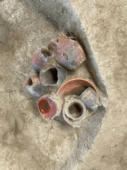 Fragmentos y pequeñas vasijas de cerámica rojiza halladas en el suelo durante una excavación arqueológica.
