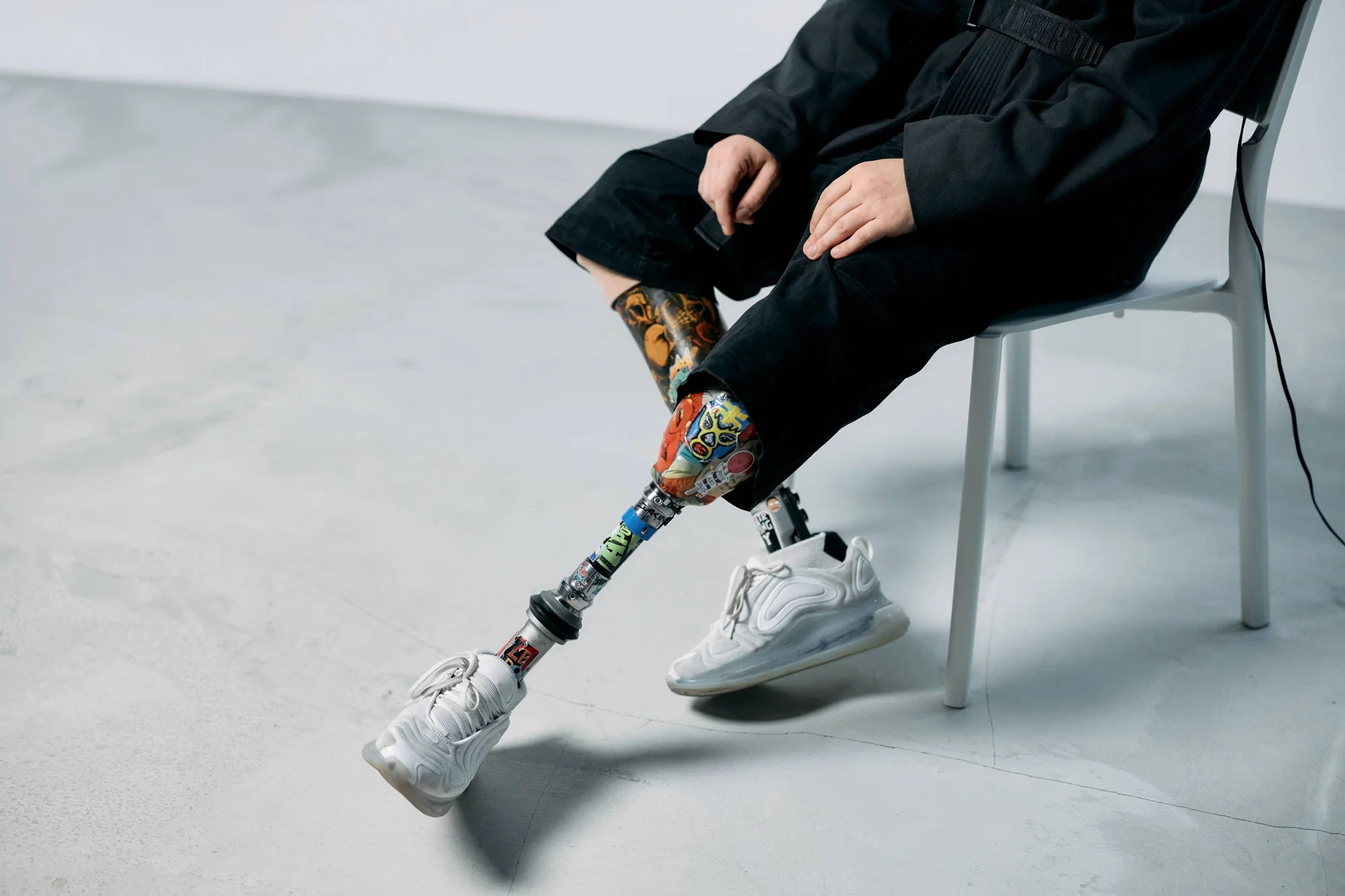 Persona sentada muestra una prótesis de pierna decorada con diseños coloridos y zapatillas blancas, junto a su pierna con tatuajes.