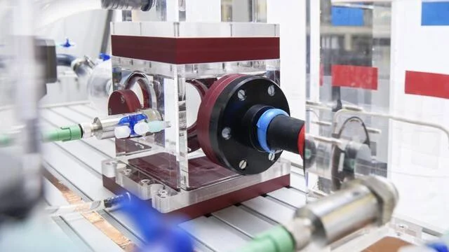 Dispositivo técnico de carcasa acrílica con conexiones de tuberías azules y verdes montado en equipo de laboratorio industrial.
