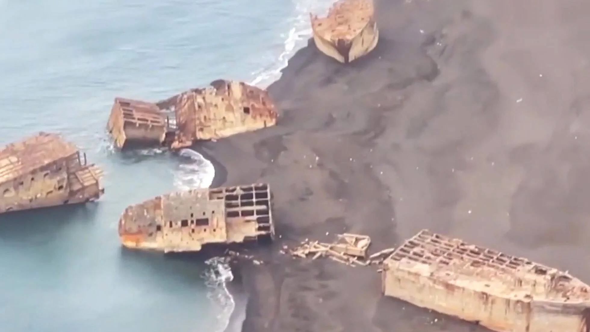 Vista aérea de varios cascos de barcos oxidados y destruidos varados en una playa de arena volcánica negra junto al mar.