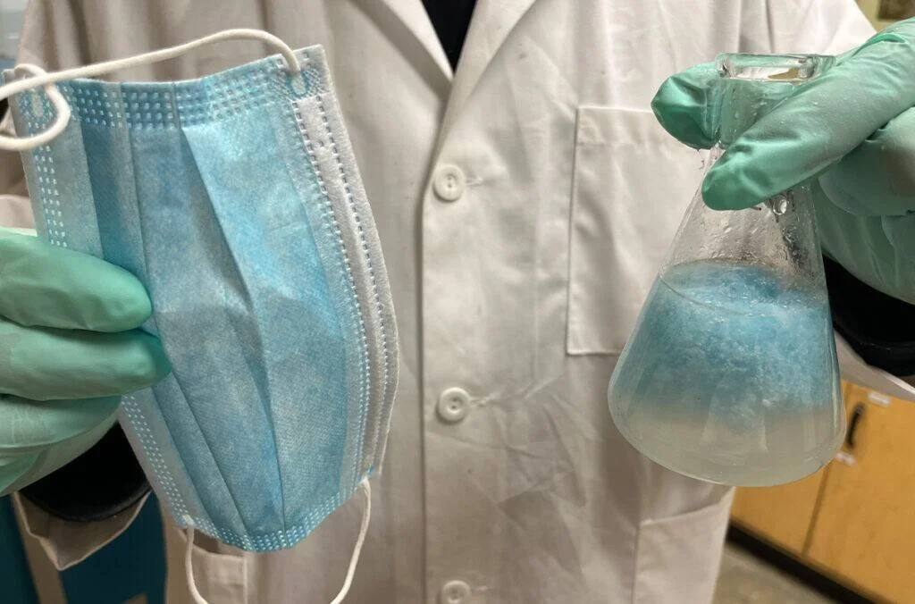 Investigador con guantes sostiene una mascarilla quirúrgica azul y un matraz Erlenmeyer con partículas azules en suspensión.