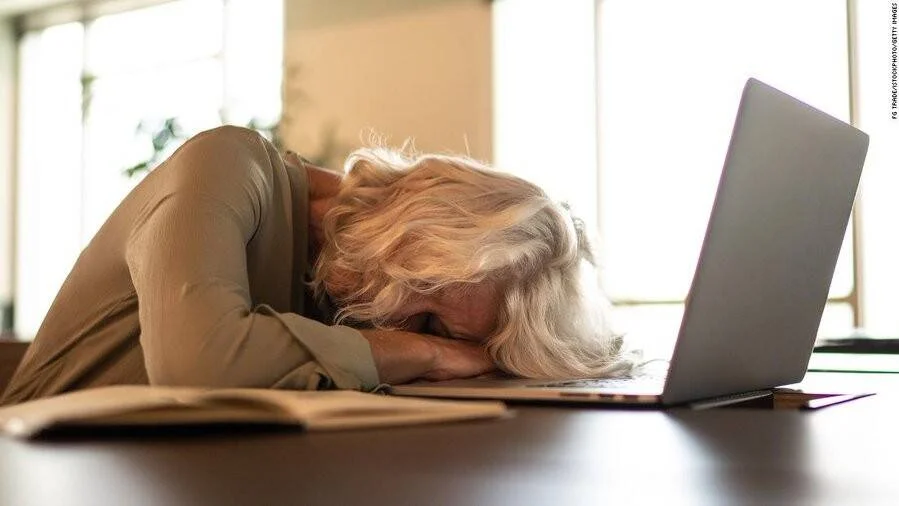 Mujer mayor con cabello blanco apoya la cabeza sobre el escritorio, exhausta, junto a una laptop y un cuaderno abierto.