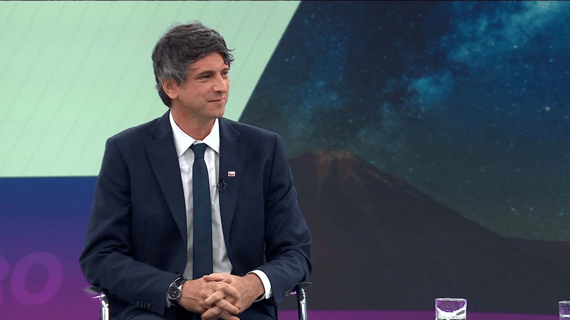 Hombre de traje azul marino y corbata oscura sentado en un estudio de televisión con fondo de volcán y cielo estrellado.