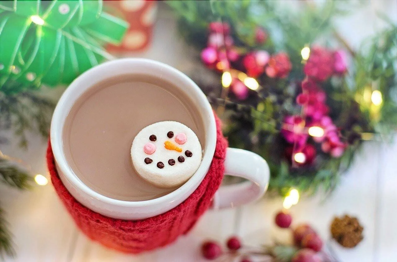 Taza de cacao caliente con funda roja de lana y un malvavisco decorado como muñeco de nieve, rodeada de adornos navideños.