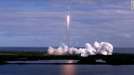 Cohete SpaceX despega dejando una gran nube de humo blanco desde una plataforma de lanzamiento junto al océano.