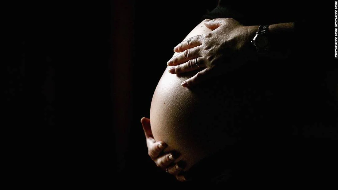 Dos manos sostienen y acarician el vientre abultado de una mujer embarazada sobre fondo oscuro.