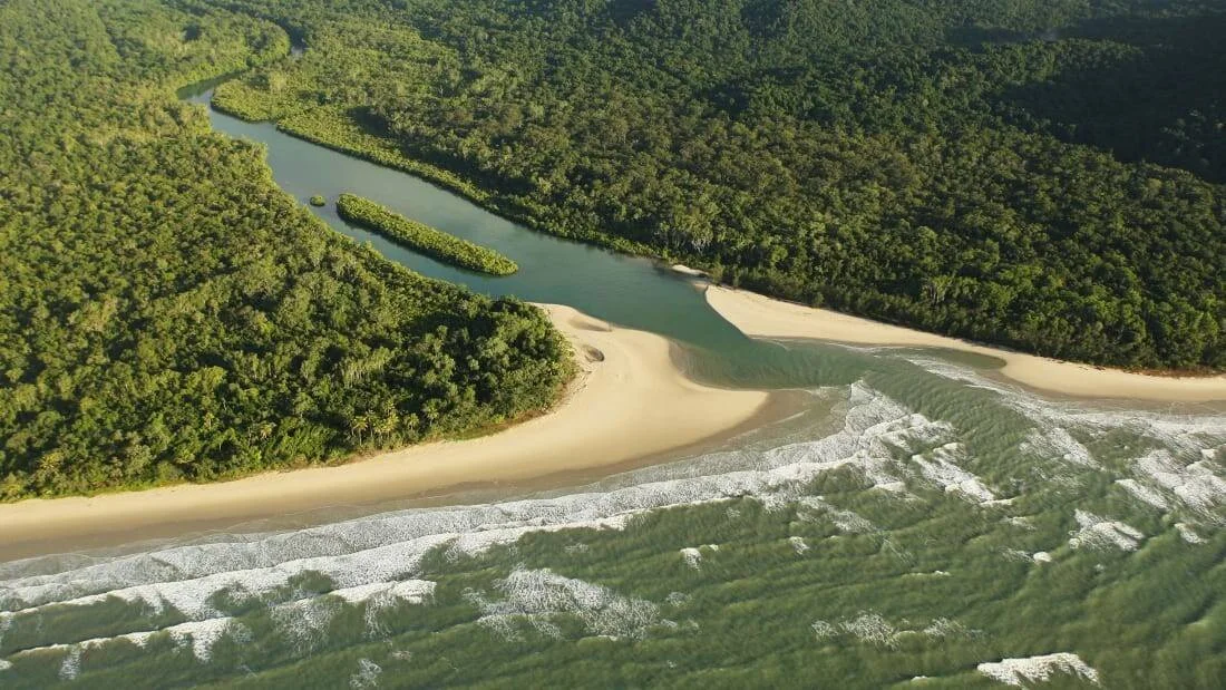 Vista aérea de un río serpenteante rodeado de selva tropical que desemboca en una playa de arena con oleaje marino.