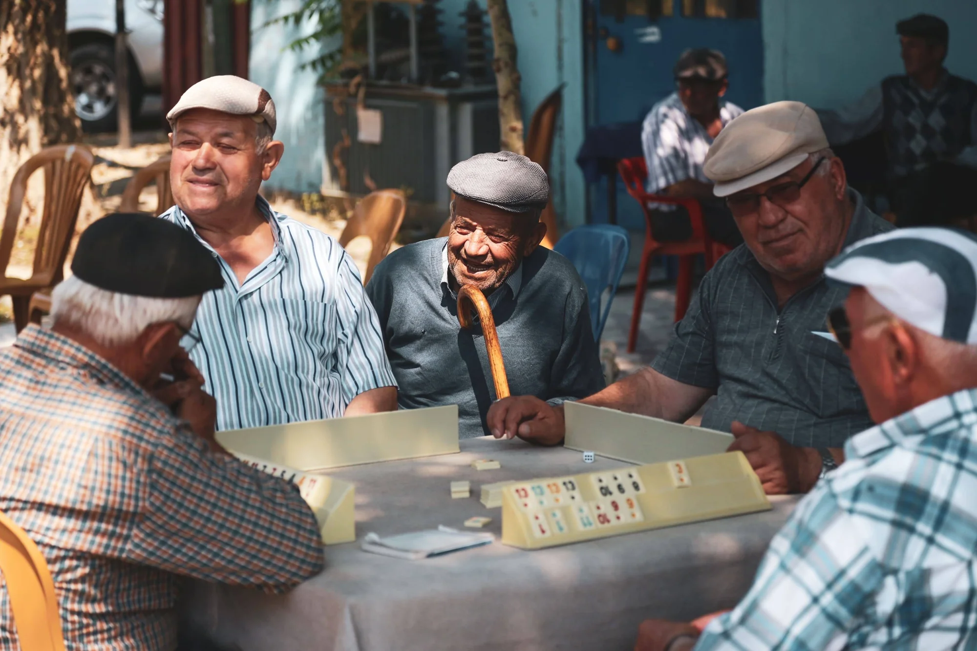 Un grupo de hombres mayores ríe y juega al dominó sentados alrededor de una mesa en un espacio exterior.