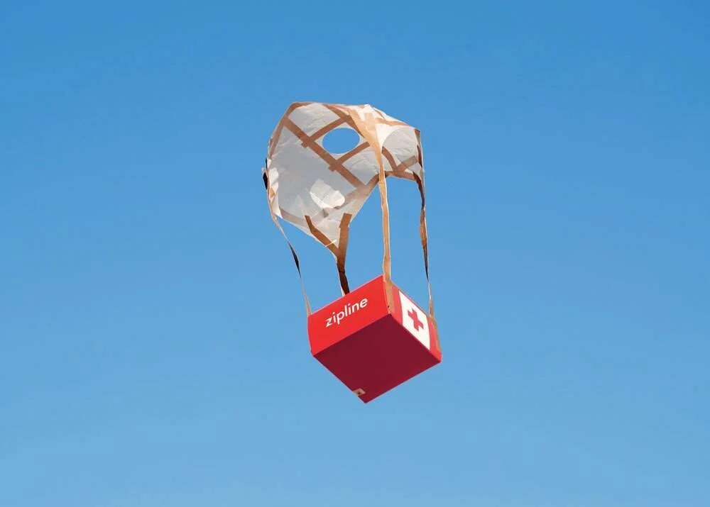 Caja roja con logo de Zipline y cruz médica desciende desde el cielo sujeta a un paracaídas de tela.