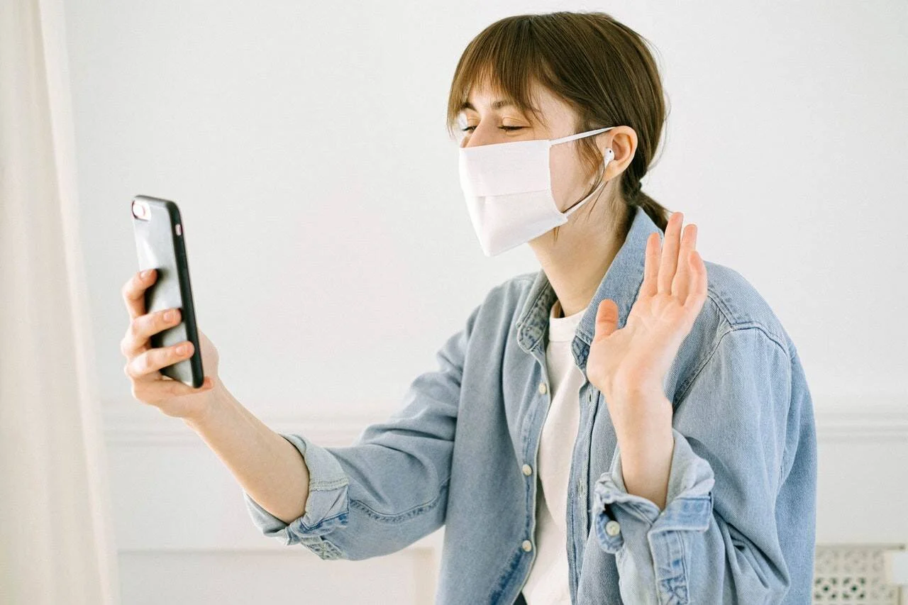 Mujer joven con mascarilla y auriculares saluda con la mano durante una videollamada desde su smartphone.