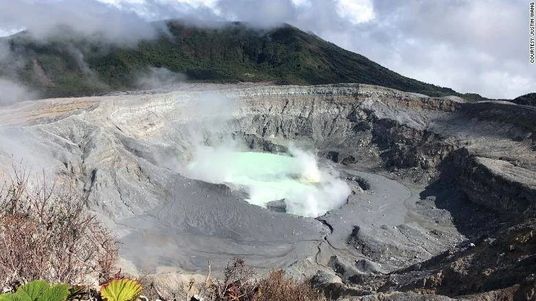 Cráter de volcán con lago de aguas ácidas de color turquesa y emisiones de gases, rodeado de paredes rocosas y vegetación.
