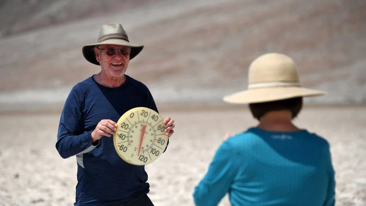 Un hombre con sombrero y gafas muestra un termómetro redondo que marca temperaturas extremas en un paisaje árido y seco.