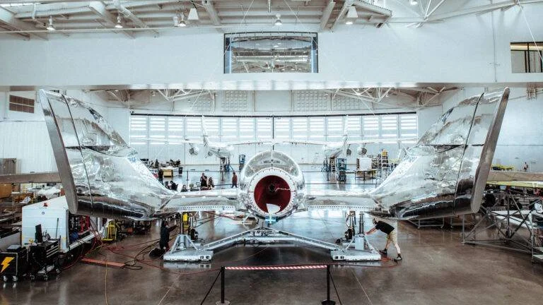 Aeronave experimental de superficie metálica pulida en proceso de ensamblaje dentro de un hangar industrial con técnicos trabajando.