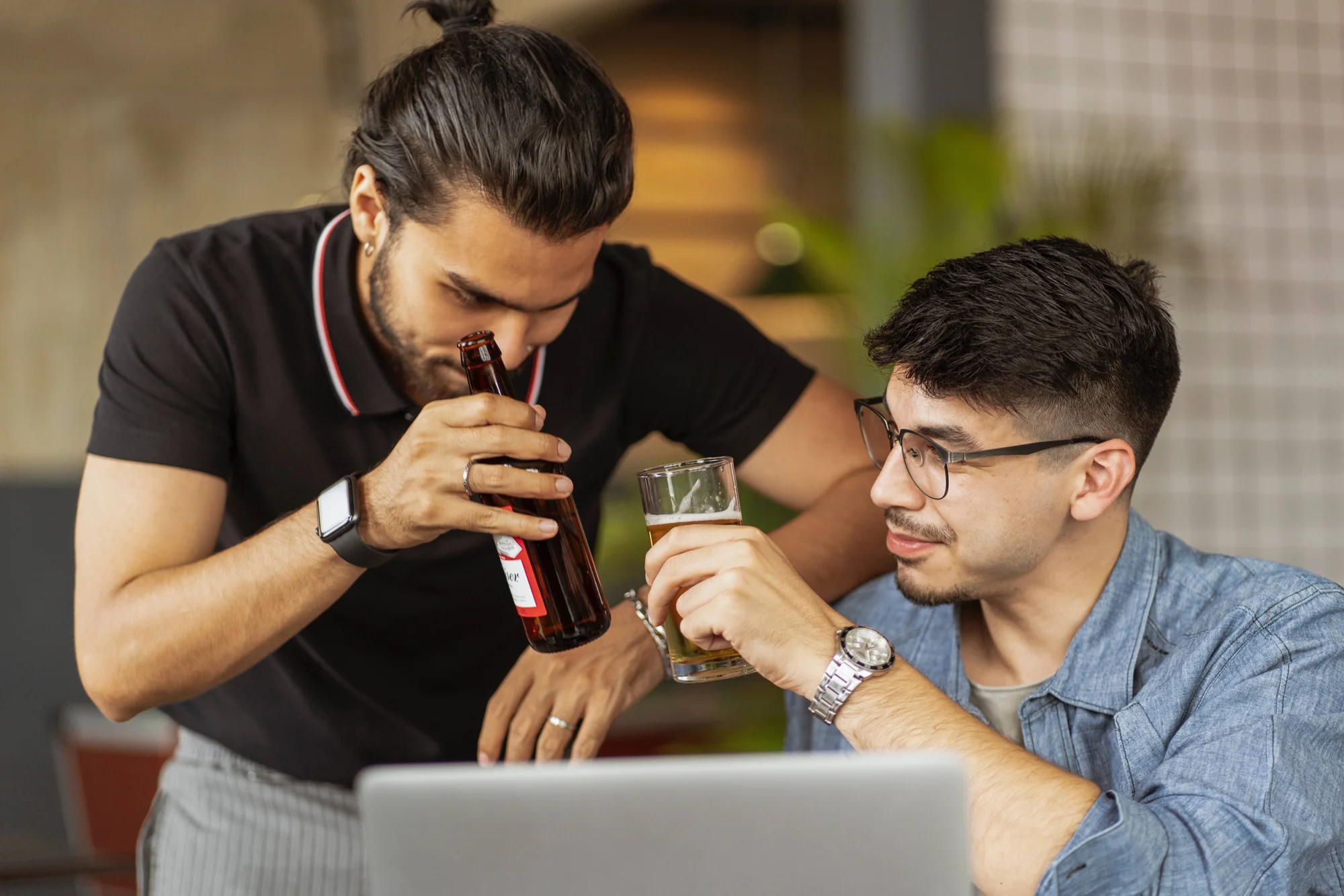 Dos jóvenes sostienen cervezas mientras miran juntos una pantalla de laptop en un ambiente informal.