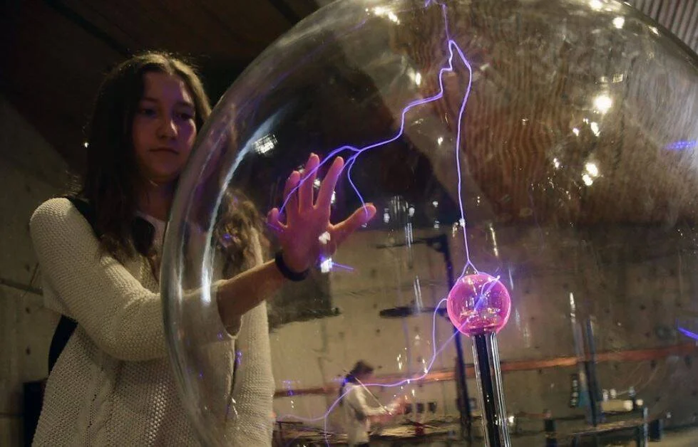 Una joven toca con la mano una gran esfera de plasma, generando rayos eléctricos violetas visibles en su interior.