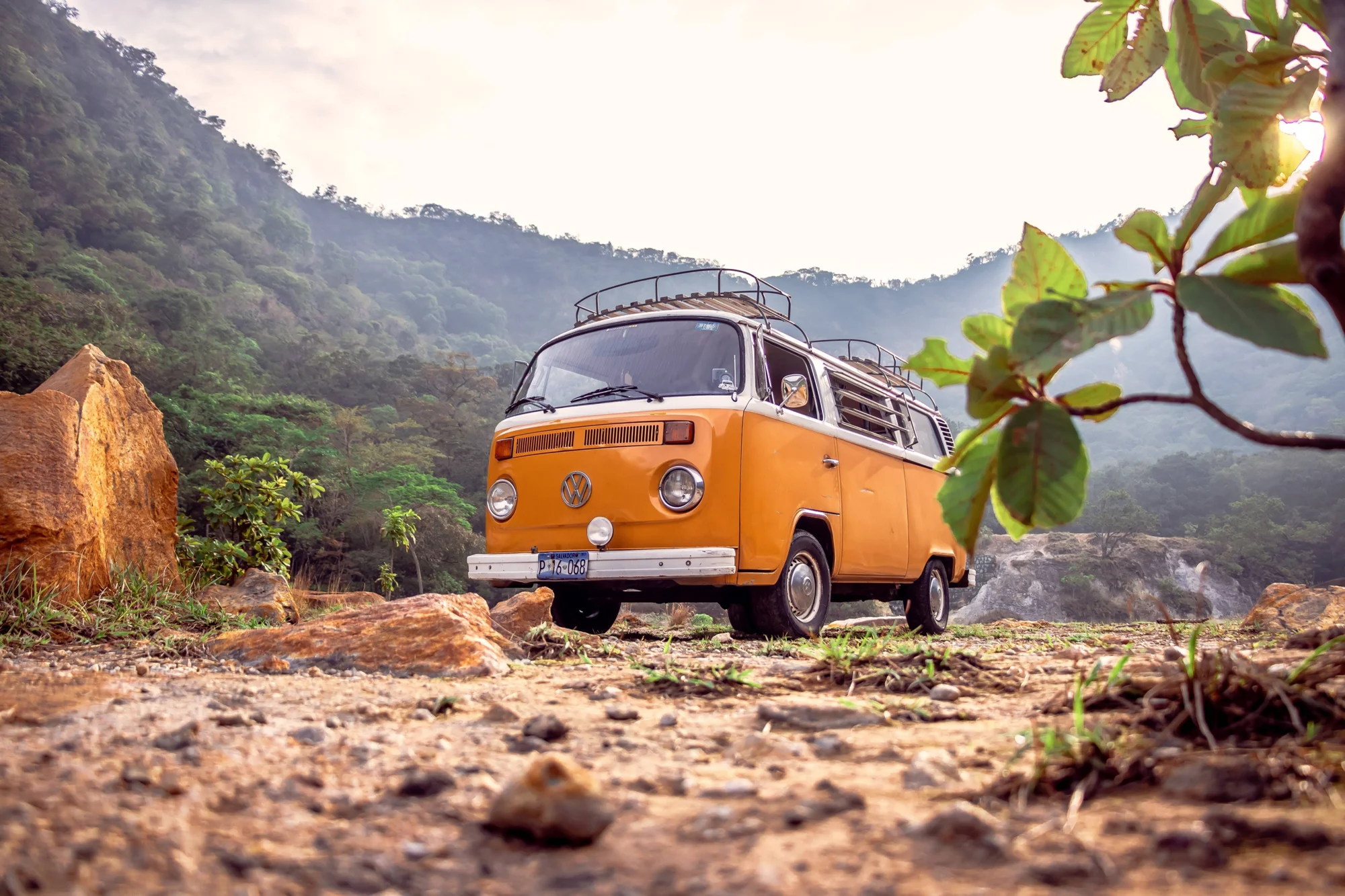 Furgoneta clásica Volkswagen Type 2 de color naranja estacionada en terreno rocoso con montañas boscosas de fondo.
