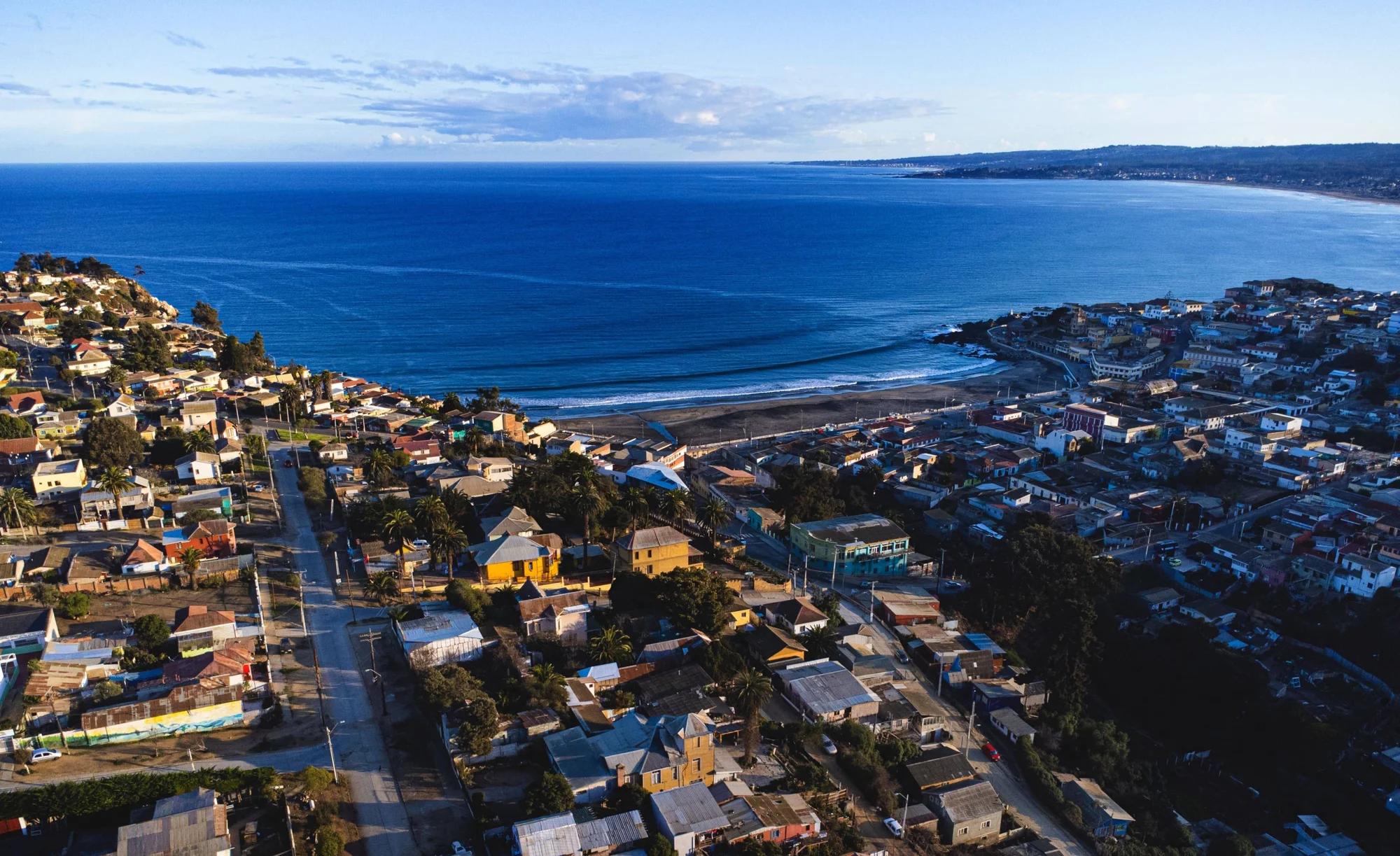 Vista aérea de una ciudad costera con casas coloridas, calles en cuadrícula y una amplia bahía de aguas azules al fondo.
