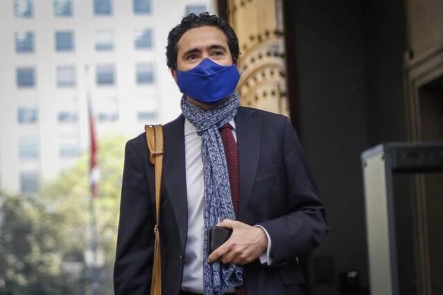 Hombre con mascarilla azul, bufanda y traje oscuro camina frente a un edificio en una ciudad.